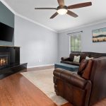 A cozy living room with wood floors, a brown leather sectional sofa, a fireplace with a mounted TV above it, a ceiling fan, and two windows with blinds letting in natural light. A landscape painting hangs on the wall.