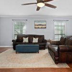 A living room with brown leather sofas, a teal ottoman, a large rug, two windows, a ceiling fan, wall art, and a small desk with a computer in the corner near the door.