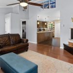 A cozy living room with brown leather sofas, a blue ottoman, a fireplace with a TV above it, and a small desk. The kitchen with wood cabinets is visible through an open doorway. Light walls and wooden floors.