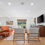 Bright living room with wood floors, orange sofa, two gray chairs, coffee table, houseplants, TV on a wooden cabinet, large windows with bamboo shades, and dining area visible in the background.