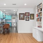 Bright, modern interior with wooden floors, a built-in white desk and bookshelf, framed art on the wall, and an open view into a turquoise-accented kitchen with bar seating and woven stools.