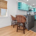 Modern kitchen with turquoise cabinets, a tiled backsplash, and a black island featuring patterned tiles. Two wicker bar stools sit by the island, and a window with a bamboo shade is on the left. The floor is wood and tile.