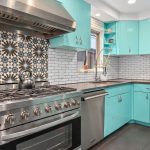 A modern kitchen with turquoise cabinets, stainless steel appliances, a patterned tile backsplash behind the stove, white subway tiles, and a window above the sink letting in natural light.