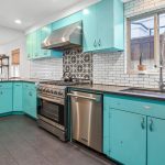 Bright kitchen with turquoise cabinets, stainless steel appliances, a large gas stove, white subway tile backsplash, patterned tile behind the stove, and a window above the sink. Light wood floors and open living area are visible.