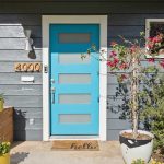 A modern house entrance with a bright blue front door, a doormat that says “hello,” a wall-mounted mailbox, potted plants, and horizontal gray siding. The house number 4000 is displayed next to the door.