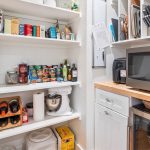 A well-organized kitchen pantry with shelves holding spices, canned goods, pasta, wine bottles, a stand mixer, paper towels, and jars; a microwave sits on a wooden countertop next to drawers.