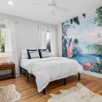 A bright bedroom with white walls and bedding, light wood floors, two small rugs, a wooden nightstand, and a feature wall mural of pink flamingos and tropical scenery. A chair and a window with sheer curtains are also visible.