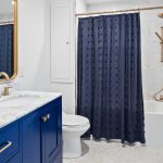 A modern bathroom with a blue vanity, marble countertop, brass fixtures, a toilet, a bathtub with a dark blue shower curtain, and blue-patterned wallpaper on one wall. Towels hang on a brass rack.