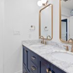 A modern bathroom with a double sink vanity, marble countertop, gold fixtures, and two mirrors. The walls are white, with a blue patterned accent wall visible in the background. A hallway with a colorful rug is also visible.