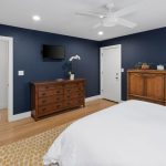 A bedroom with dark blue walls, light wood floor, a white bedspread, wooden dresser and cabinet, wall-mounted TV, ceiling fan, and some potted plants and decor on the furniture.
