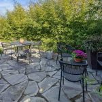 Outdoor patio with stone flooring, surrounded by lush green bamboo. Black metal tables and chairs are arranged with potted purple flowers on the tables, creating a cozy, inviting garden seating area.