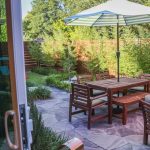 A backyard patio with wooden furniture and a table under a green-and-white striped umbrella, surrounded by lush plants and a wooden fence, viewed from an open glass door.