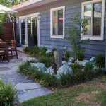 A backyard with a stone patio, wooden outdoor dining set, umbrellas, and a small landscaped garden with stones and greenery next to a gray house with multiple windows.