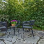 Two black chairs and a small round table with a potted purple flower sit on a stone patio, surrounded by lush green bamboo plants and garden vegetation.