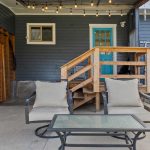 A cozy backyard patio features two cushioned chairs and a glass-top table, set on concrete near wooden stairs leading to a blue door. String lights hang above, and wooden fencing surrounds the area.