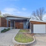 A modern single-story house with a slanted roof, wooden fencing, and a one-car garage. The driveway splits into two paths, with a small grassy area and scattered leaves in front. Trees are visible in the background.