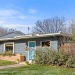 A small, single-story blue house with a bright turquoise front door and large windows sits next to a driveway. The yard has green grass, shrubs, and some trees in the background under a blue sky.