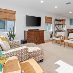 A bright living room with patterned furniture, a wooden cabinet under a wall-mounted TV, two chairs, potted plants, and a view into a kitchen with teal cabinets and a dining area.