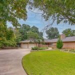 A single-story brick house with a large curved driveway, two-car garage, well-kept lawn, garden beds, and tall green trees on a cloudy day.