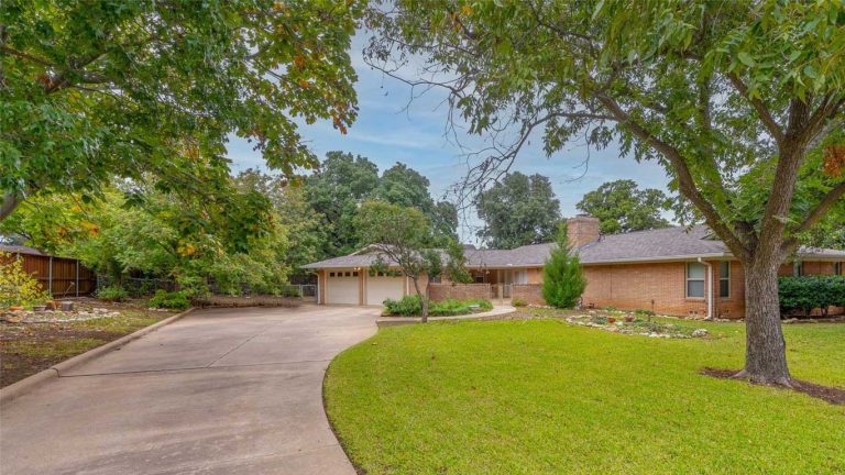 A single-story brick house with a large curved driveway, two-car garage, well-kept lawn, garden beds, and tall green trees on a cloudy day.