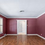 An empty room with maroon walls, white crown molding, and wooden floors. A large window with white shutters is on the left, and double doors open to another brightly lit space.