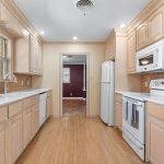 Bright kitchen with light wood cabinets, white appliances (refrigerator, stove, microwave, dishwasher), tile backsplash, white countertops, and wood flooring; view into adjoining room with maroon walls.