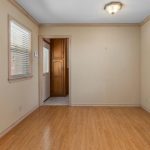 A small, empty room with light wood flooring, beige walls, white trim, two windows with blinds, and a doorway leading to another room. A ceiling light fixture is centered on the white ceiling.