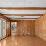 A spacious room with wood-paneled walls, wood flooring, a white drop ceiling with exposed wooden beams, and double glass doors on one side. There are electrical outlets and cords visible along the walls.