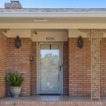 A brick house front porch with a glass storm door, house number 4216 above the door, two wall lanterns, a potted plant on the left side, and decorative trim near the door.
