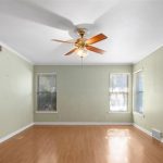 Empty room with light green walls, two windows with blinds, a ceiling fan with a light, wood-look flooring, and white crown molding. Sunlight streams through one window, creating a bright spot on the floor.