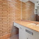 A bathroom with a brick accent wall, large mirror, white vanity, and a tan tile countertop with a built-in sink. A white door and small window with blinds are visible in the background.
