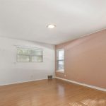 Empty room with light hardwood floors, a white ceiling, and a mix of white and pale pink walls. Two windows with white blinds let in natural light, and there is a simple square ceiling light fixture.