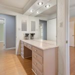 A built-in vanity with drawers and a large mirror is in a dressing area with light wood flooring, near a hallway and a bedroom with closets and a ceiling fan.