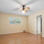 Empty room with light-colored walls, wood flooring, a ceiling fan with lights, two windows, a closet with white doors, and an open doorway leading to another room. Natural light comes through the windows.