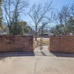 A brick patio area with a short brick wall and an iron gate, bordered by two houses. Bare trees and shrubs are visible beyond the gate under a clear blue sky.