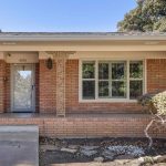 A brick house with a covered front porch, decorative columns, a glass door with the number 4216, potted plant by the entrance, large window with white shutters, and trimmed bushes along the edge.