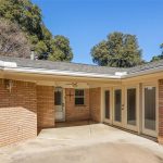 A private, paved patio area bordered by tan brick walls and tall windows with white trim. There are two light fixtures, a ceiling fan, and mature trees visible beyond the roofline under a clear blue sky.