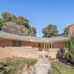 Single-story brick house with a gray shingle roof, attached two-car garage, gated entryway, and a small tree in the front yard on a sunny day.