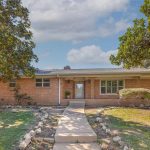 Single-story brick house with a central pathway leading to the front door, surrounded by large trees and a grassy yard under a partly cloudy sky. Shrubs and plants line the walkway and the house’s foundation.