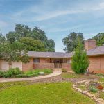 Single-story brick house with a curved driveway, a small front garden with rocks and potted plants, and mature trees in the background under a partly cloudy sky.