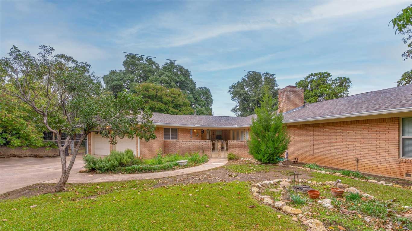 Single-story brick house with a curved driveway, a small front garden with rocks and potted plants, and mature trees in the background under a partly cloudy sky.