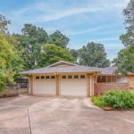 Single-story brick house with two-car garage, surrounded by trees and greenery. Wide driveway leads to the garage; the yard features shrubs and a curved walkway near the front porch. Sky is partly cloudy.