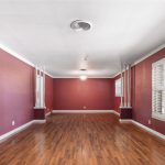 A spacious, empty room with reddish-purple walls, wood floors, white crown molding, and large windows with white shutters letting in natural light. Two ceiling lights are visible, and pillars accent the corners.