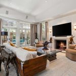 Spacious living room with large windows, neutral-toned furniture, a fireplace with a TV above it, and art sculptures. Light streams in through glass doors leading to a backyard, with beige curtains framing the view.