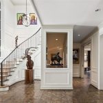 A bright, elegant foyer with a curved staircase, large windows, art pieces on the walls, sculptures on pedestals, and wood floors. Built-in shelves and rooms are visible through open doorways.