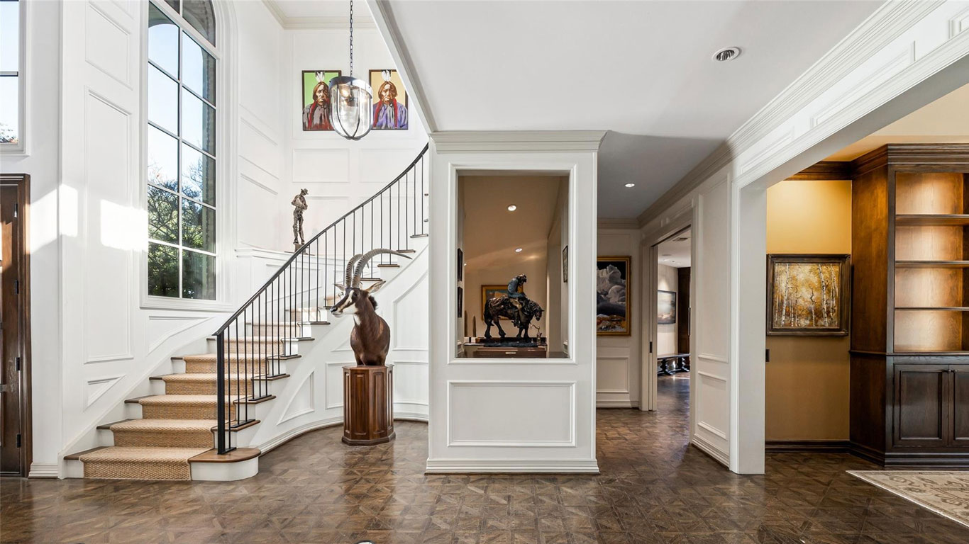 A bright, elegant foyer with a curved staircase, large windows, art pieces on the walls, sculptures on pedestals, and wood floors. Built-in shelves and rooms are visible through open doorways.