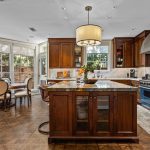 A spacious, elegant kitchen with dark wood cabinets, a large island with barstools, stainless steel appliances, marble countertops, and a dining area by large windows overlooking a brick wall and greenery.