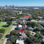 Aerial view of a suburban neighborhood with many trees, a golf course on the left, and a red location pin marking a house in the center; city skyline visible in the distance under a clear blue sky.