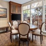 A dining area with a round wooden table and four upholstered chairs, large windows with views of a brick wall and trees, a TV on the wall, and a bench beneath a landscape painting.