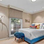A cozy bedroom features a bed with colorful pillows, a blue cushioned bench, a rustic nightstand with a lamp, and a window with plaid blinds. Antlers on the wall cast a dramatic shadow next to the window.